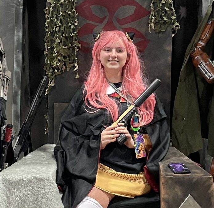 Megan in an cosplay as a teen age Anya from Spy X Family holding a pistol with a silencer while sitting on the Mandalorian throne.