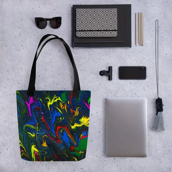 Tote bag with bright swirls of red, blue, and yellow mix with green representing the main characters on a glittering charcoal background to invoke the feel of space and black handles surrounded by accessories like sunglasses and a laptop demonstrating what can go into the bag.