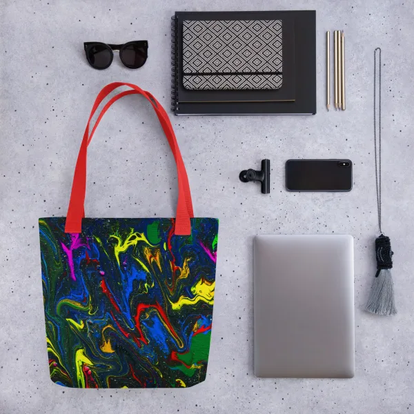 Tote bag with bright swirls of red, blue, and yellow mix with green representing the main characters on a glittering charcoal background to invoke the feel of space and red handles surrounded by accessories like sunglasses and a laptop demonstrating what can go into the bag.