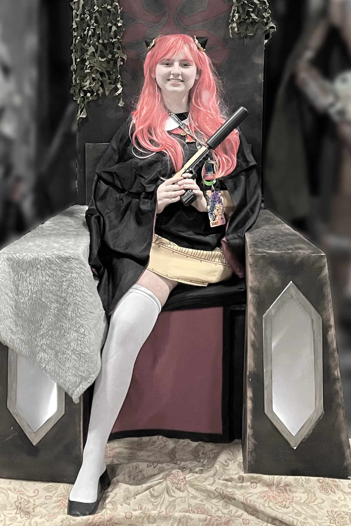 Megan in an cosplay as a teen age Anya from Spy X Family holding a pistol with a silencer while sitting on the Mandalorian throne.